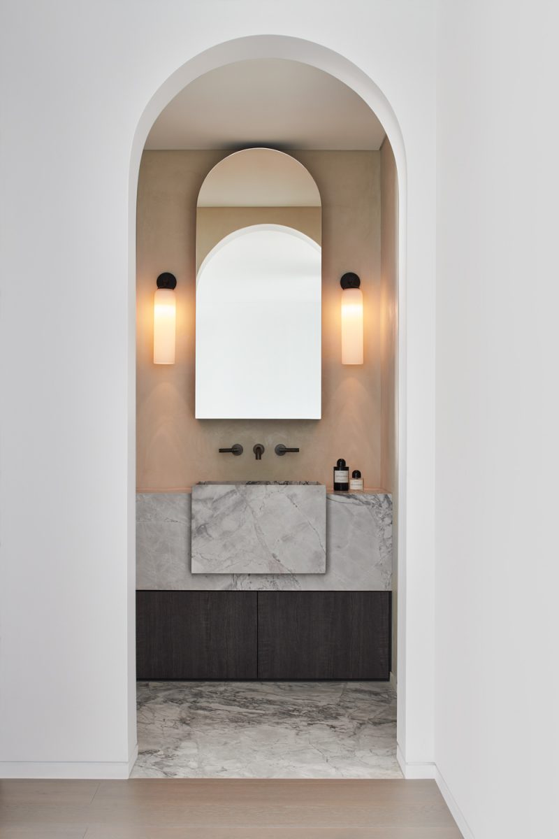 Minimalist London bathroom interior design with marble vanity, arched entryway, and ambient wall lights by The Stylesmiths.