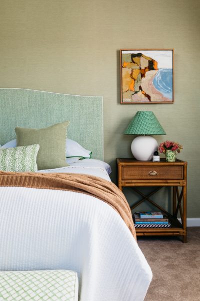 Green bedroom with patterned lamp, rattan bedside table, and abstract artwork designed by The Stylesmiths London.