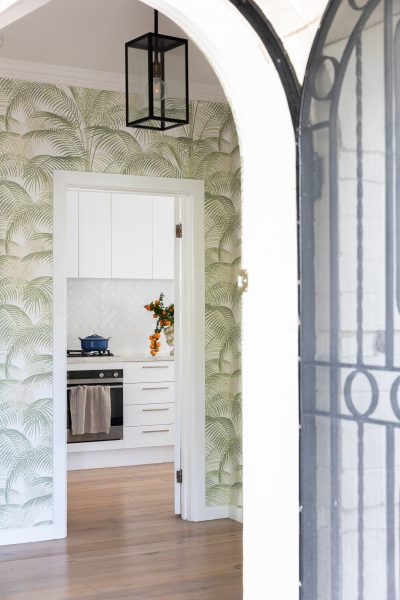 Arched doorway leading to a white kitchen with herringbone tiles and palm wallpaper, designed by The Stylesmiths London.