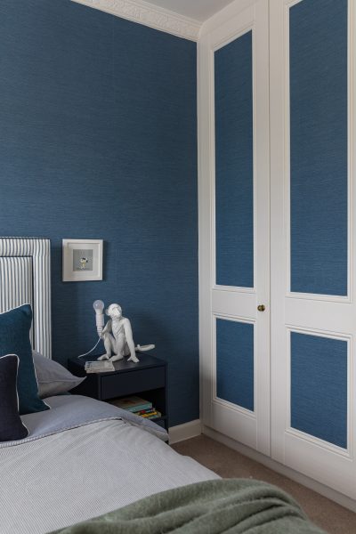 Blue wallpapered child’s bedroom with white panelled wardrobe and modern bedside table.